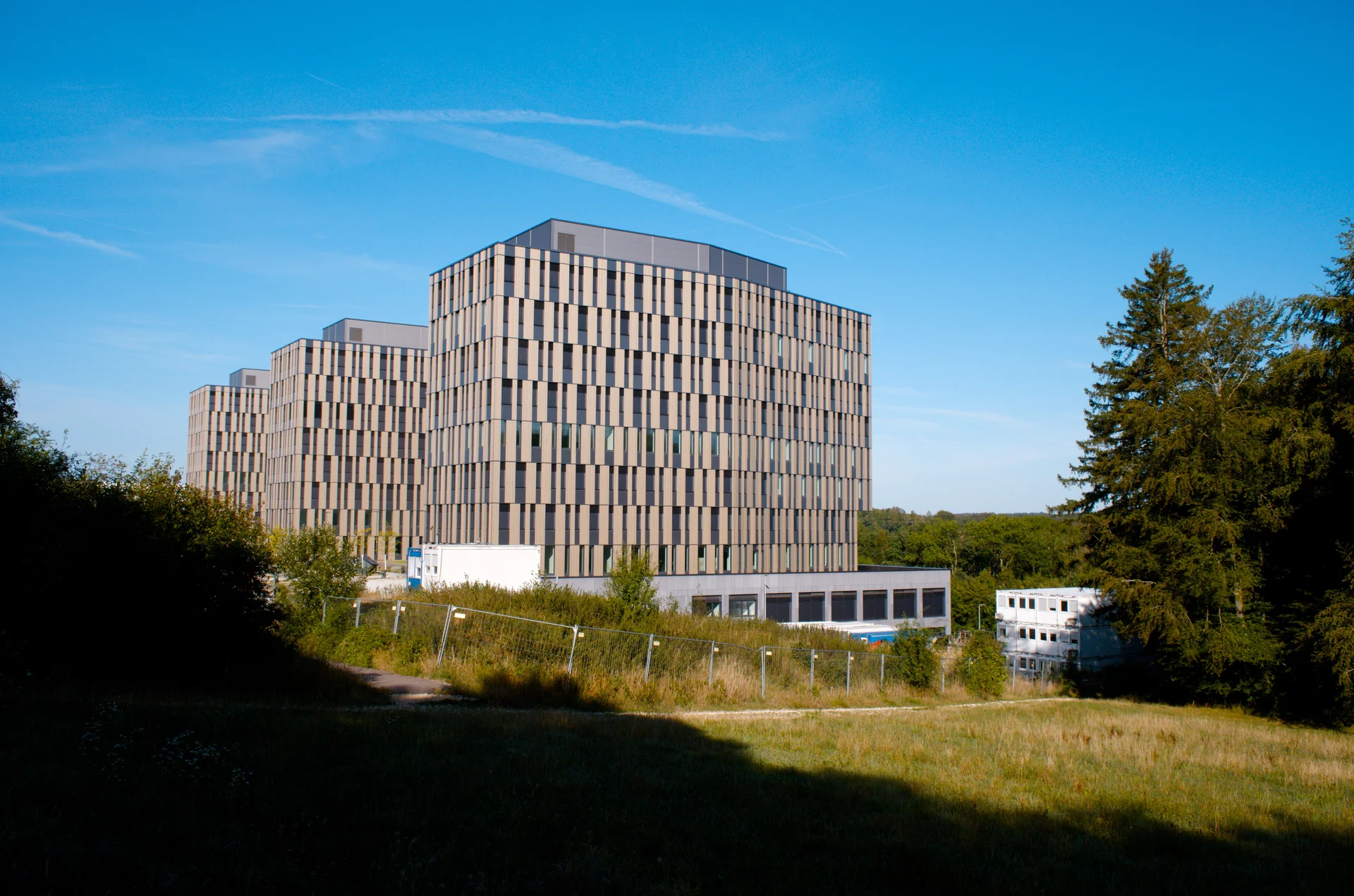 A large and very new office building besides a woodland. Next to the building is a comparably very small three-story container complex as a construction office.