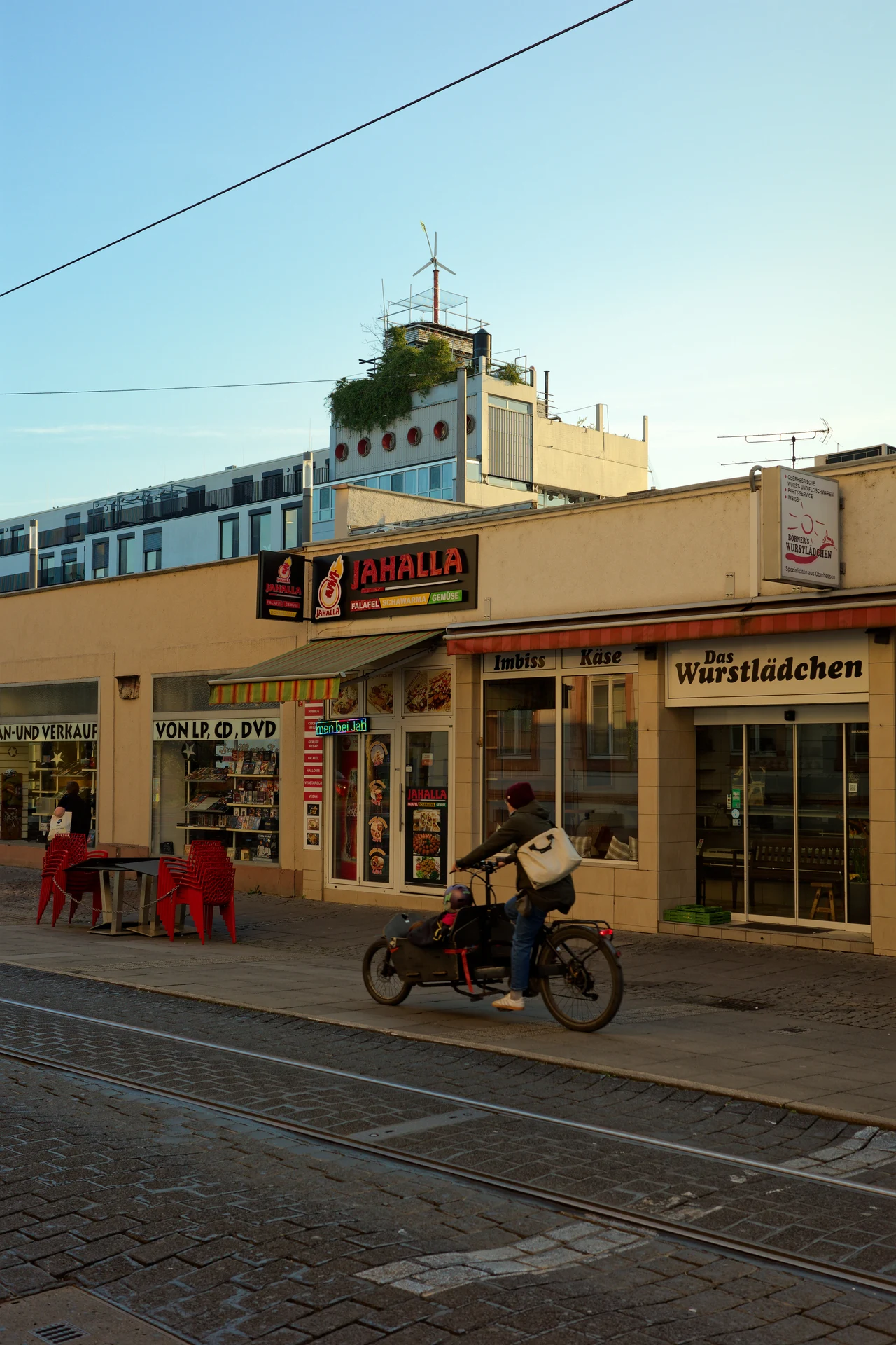 A person with a cargo bike is driving in front of a few stores in a low building. In the background, a larger building with a wind wheel on top.
