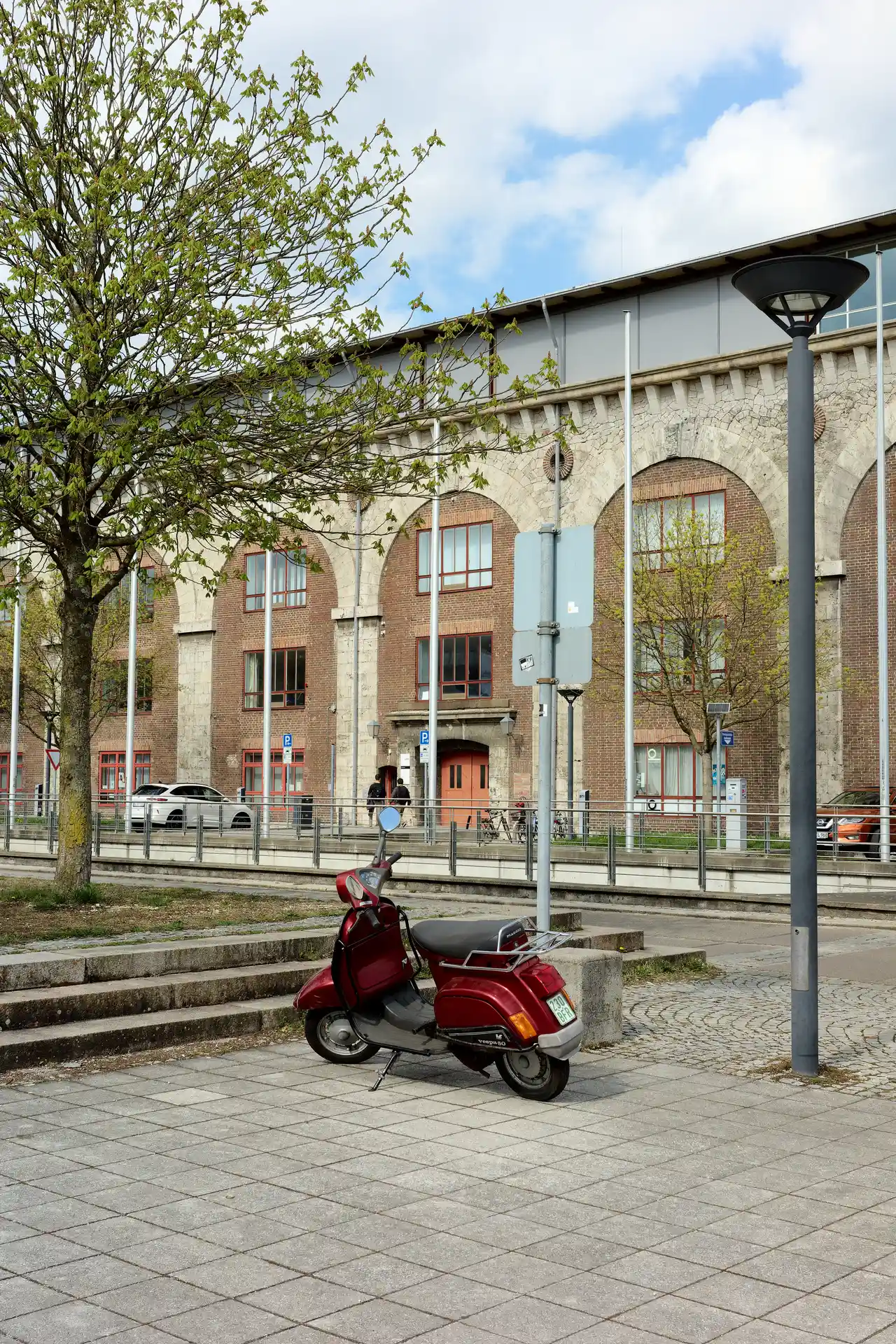 An older, dark red, classical scooter is parked in the corner of a square. In the background is a historic building, and on the left is a tree with fresh leaves.