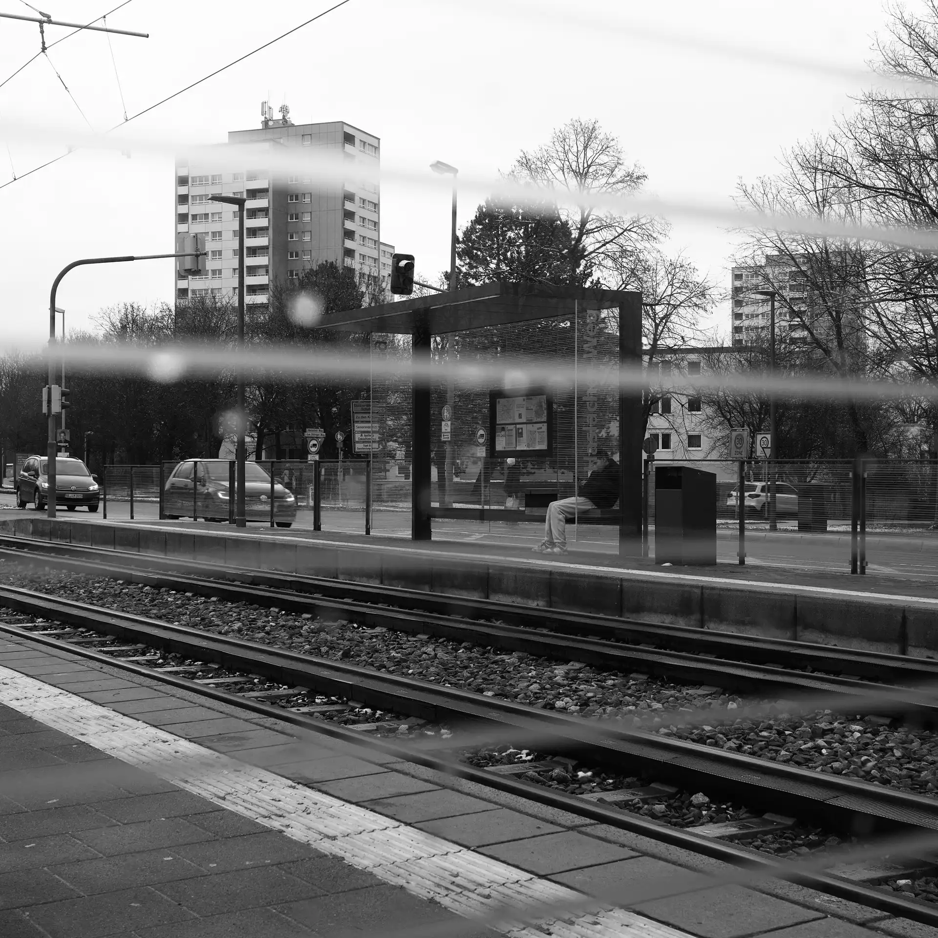 A person waiting at the opposite waiting area of a tram station. Photographed through the glass of the waiting shelter which has some lines printed on it.