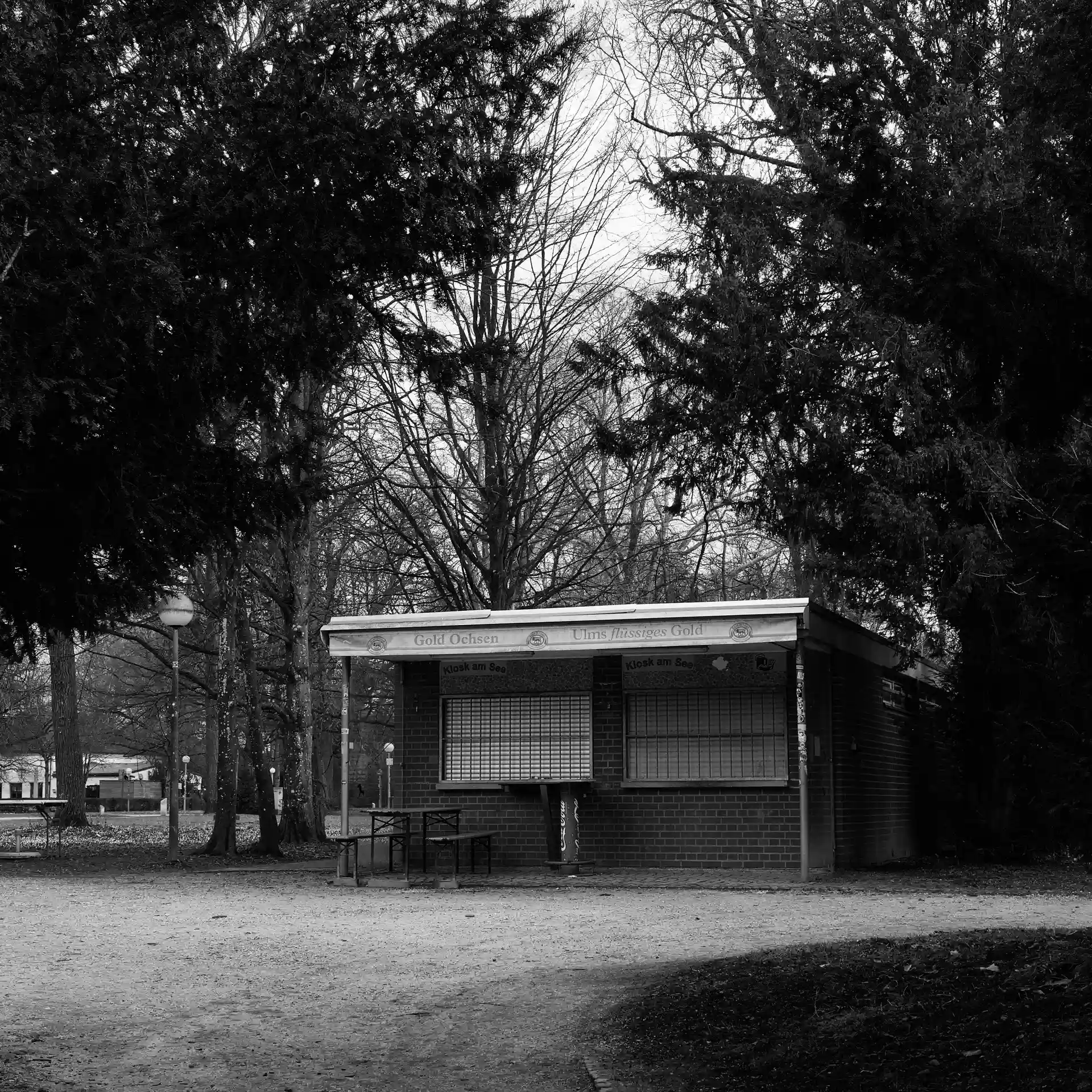 A closed kiosk building in a park with a beer garden set in front of it. Framed by trees.