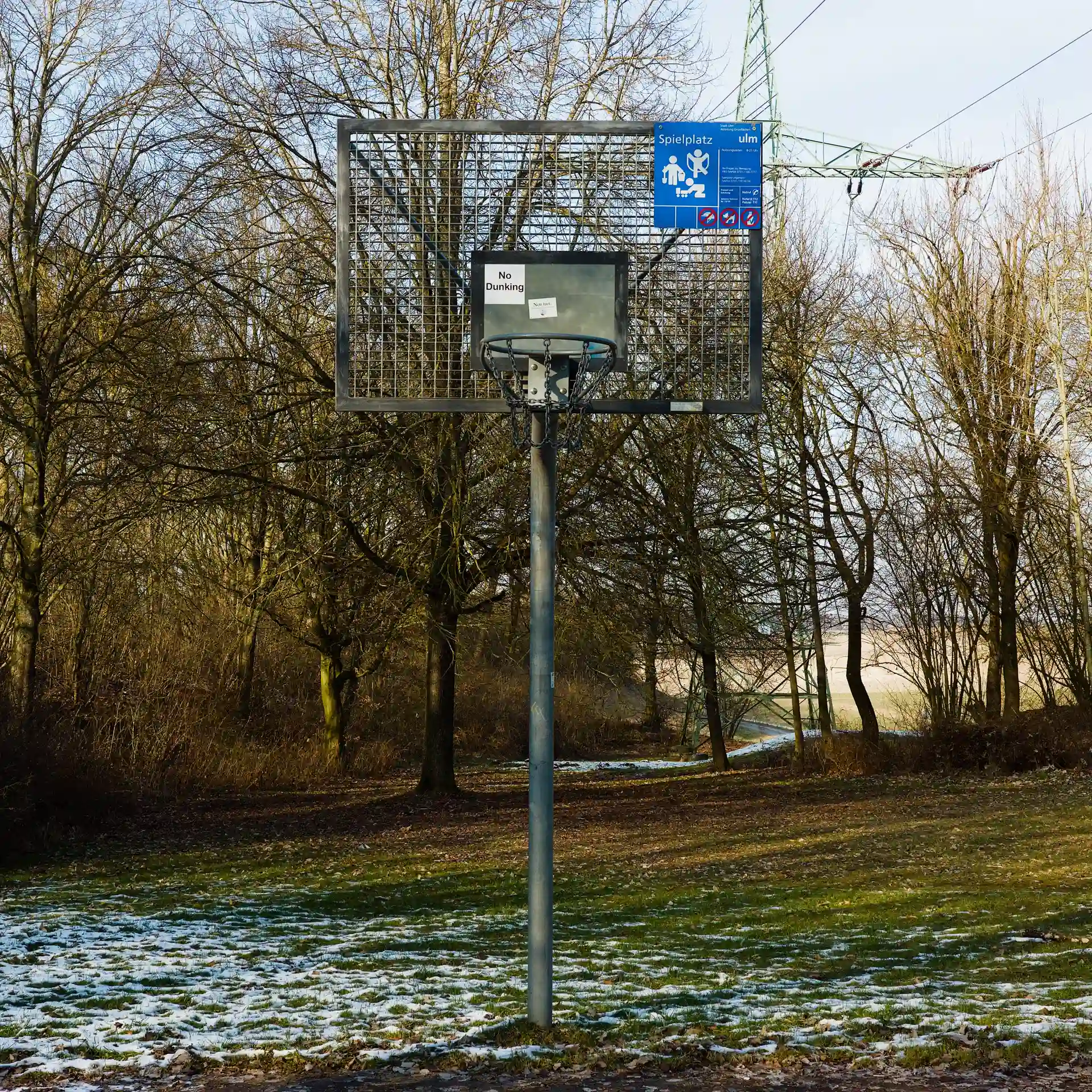 An all-metal outdoor basketball hoop in a park. A bit of snow lies on the ground.