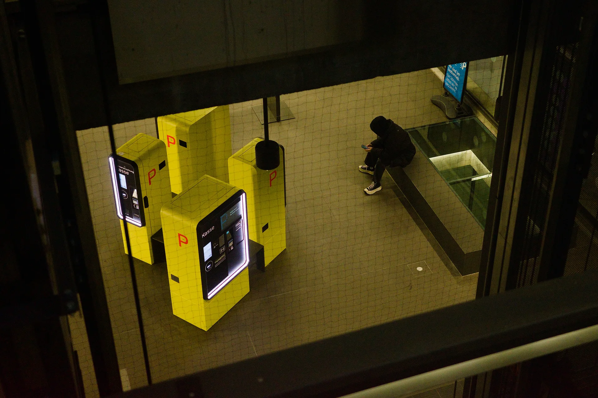 A colour photo of a subground floor photographed from above. The view is heavily framed by the architecture of the building. Four parking ticket vending machines can be seen which are painted with a vibrant yellow. A person is sitting on a bench next to the machines looking at their smartphone.
