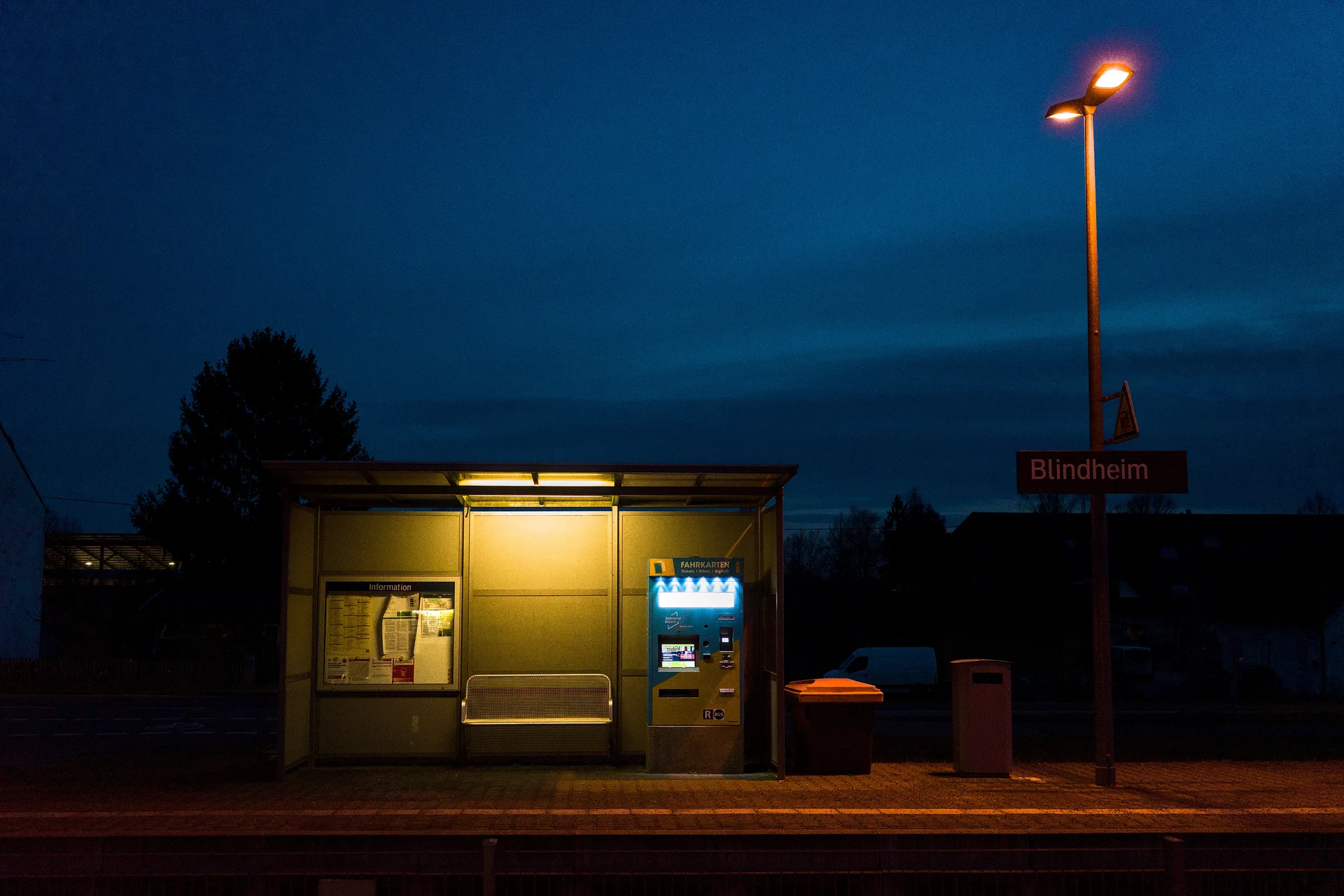 A colour photo during late sunset of a waiting shelter at a train platform. The shelter is on the left and middle of the frame and on the right is a street lamp. The differnt colour temperatures of the sky, the street lamp and the lamp in the shelter work very well together.