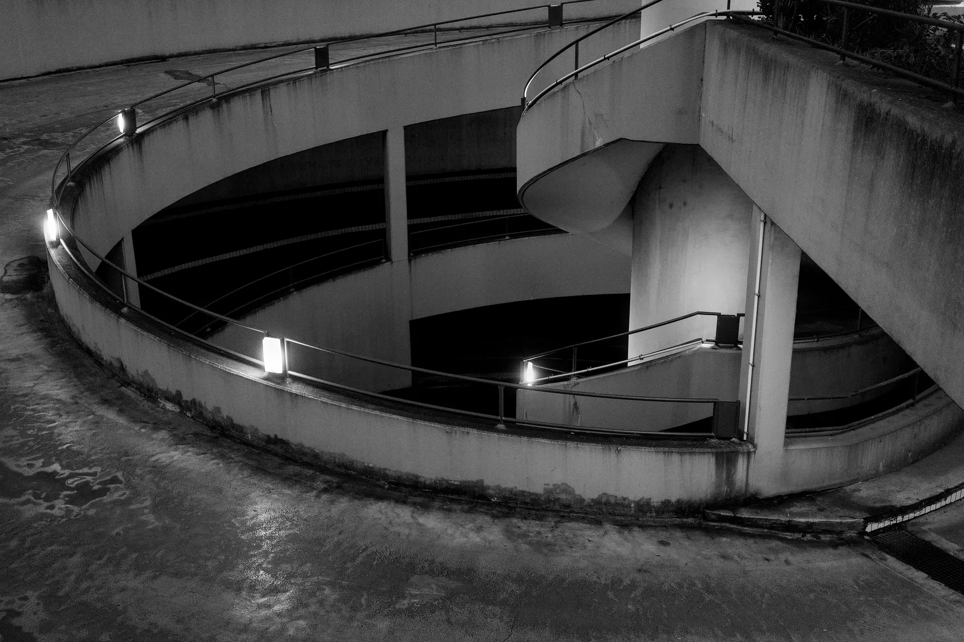 A black and white photo of a car entrance to an underground carpark during evening. The architecture is perhaps from the 70s or 80s with a lot of visible concrete. A part of the spiral road can be seen in the entire fram and on the top right corner is a part of the pedestrian access. The area is lit by lights build into the railing.