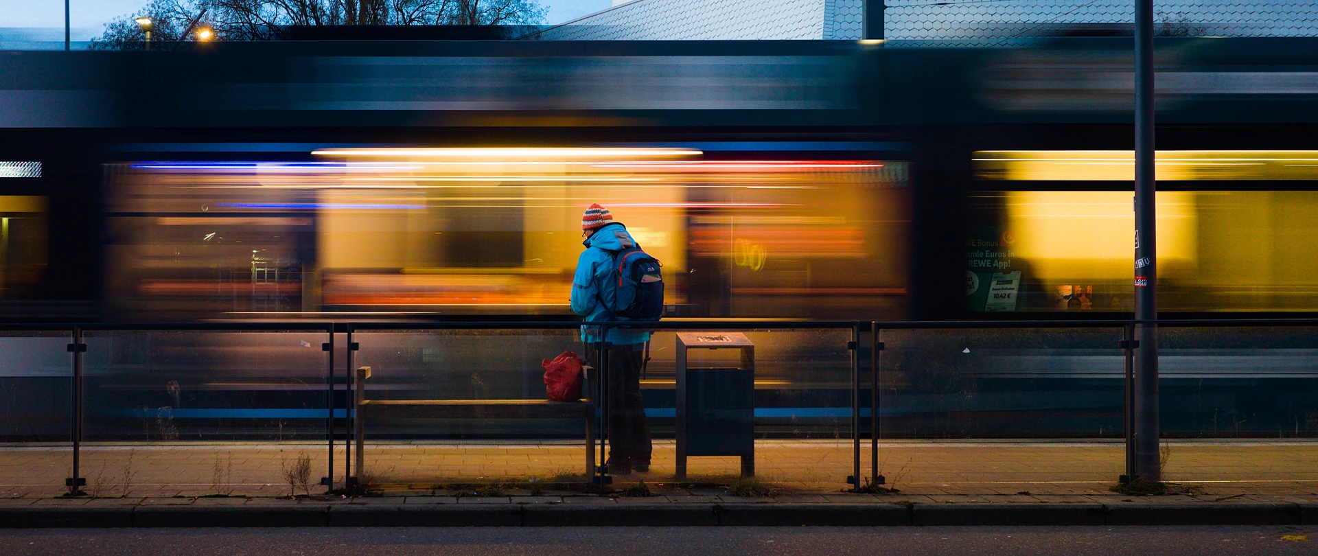 A long-exposure colour photo in the evening. In the background a tram passing by. Due to the long exposure the lights of the tram are smushed horizontally with a brighter patch in the middle. In front of that a person is standing, wearing a bright blue jacket and with a vibrant red back next to them. The colours are very contrastful compared to the warm lights of the tram in the background.