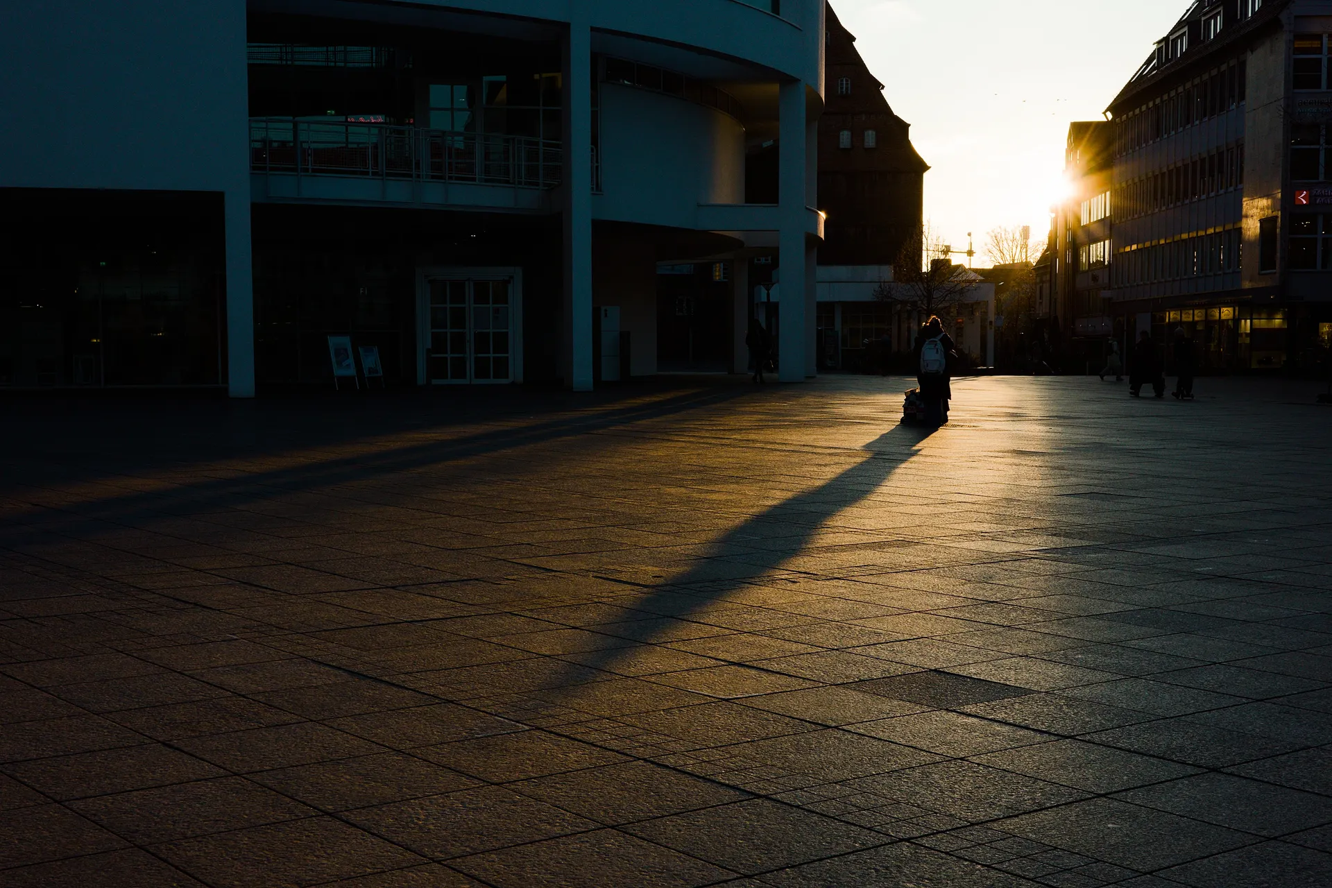 A colour photo on the minster place in Ulm. On the left and middle is the Stadthaus, on the right edge another building. Between is a gap between the buildings giving way to the descending sun which gives the whole scenery a rather warm colour. A person is standing in the middle of the beam of light and throwing a long shadow.