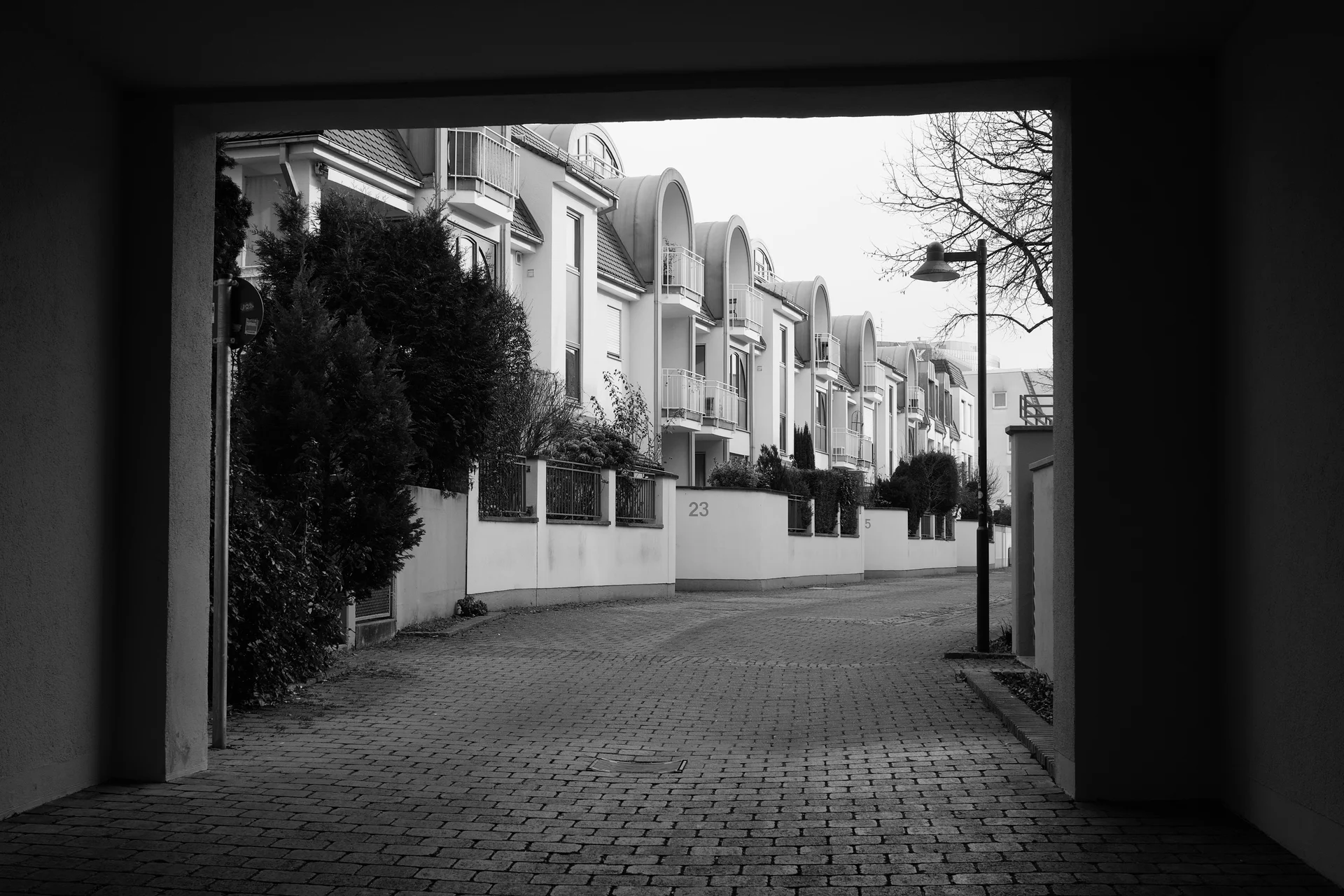 Black and white photo. The scene is framed by a rectangular archway. A row of of buildings all of the same architecture can be seen from the middle of the frame going towards the background. The buildings all have a small frontyard surrounded with brick walls.