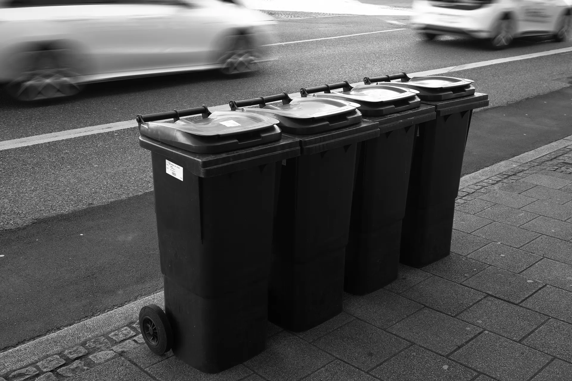 Picture of four wheelie bins in a row on a sidewalk. In the background a road with two cars partly visible. These are quite blurred due to slow shutter speed.