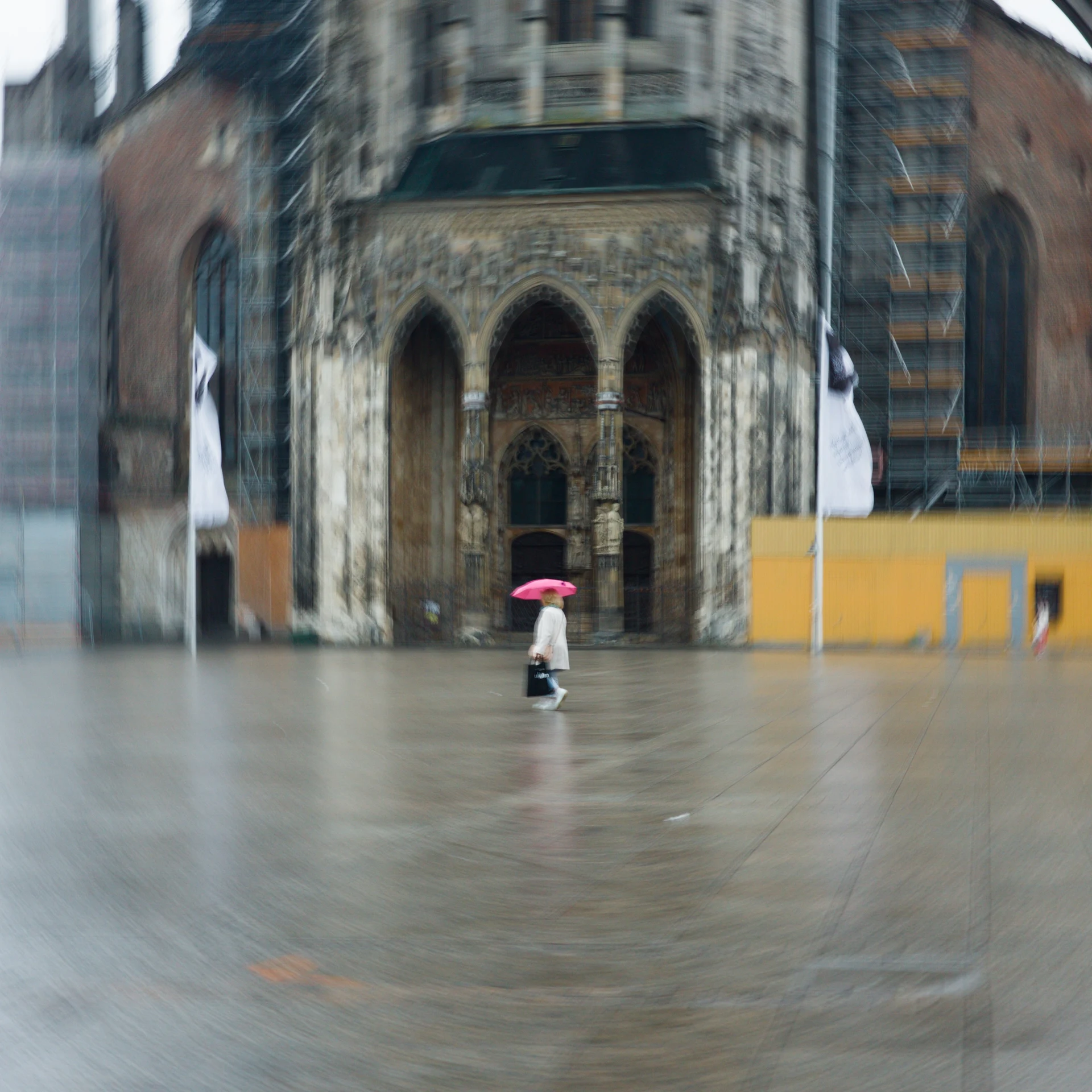 Picture of a person with a white jacket and a pink umbrella on the place in front of the Ulmer Münster during rain. The camera was rotated during exposure resulting in a circular effect.